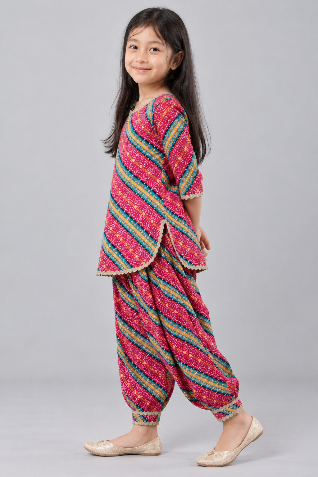 A young girl modeling a vibrant pink Patola-style two-piece kurta set with multicolored stripes and cream scalloped edges, paired with beige flat shoes, against a solid gray studio background.