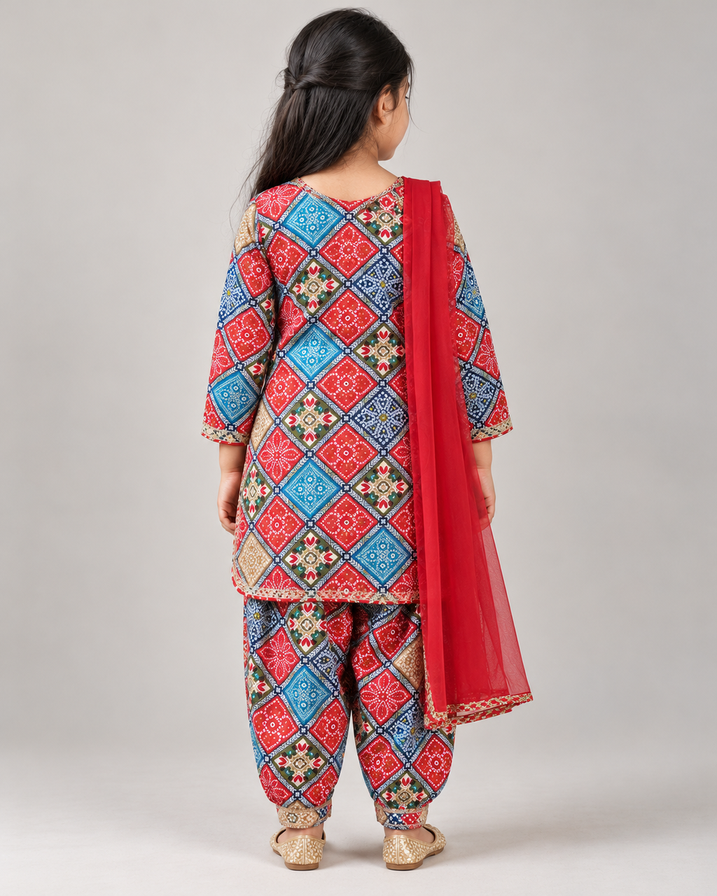 A child model shown from the back wearing a traditional Indian salwar kameez set with a red and blue geometric pattern, paired with a solid red dupatta, on a neutral gray background.
