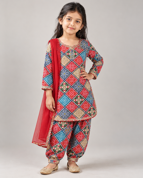 A young girl modeling a traditional Indian salwar kameez suit with a red dupatta, featuring a multicolor geometric patterned kurta and matching pants, set against a plain gray studio background.